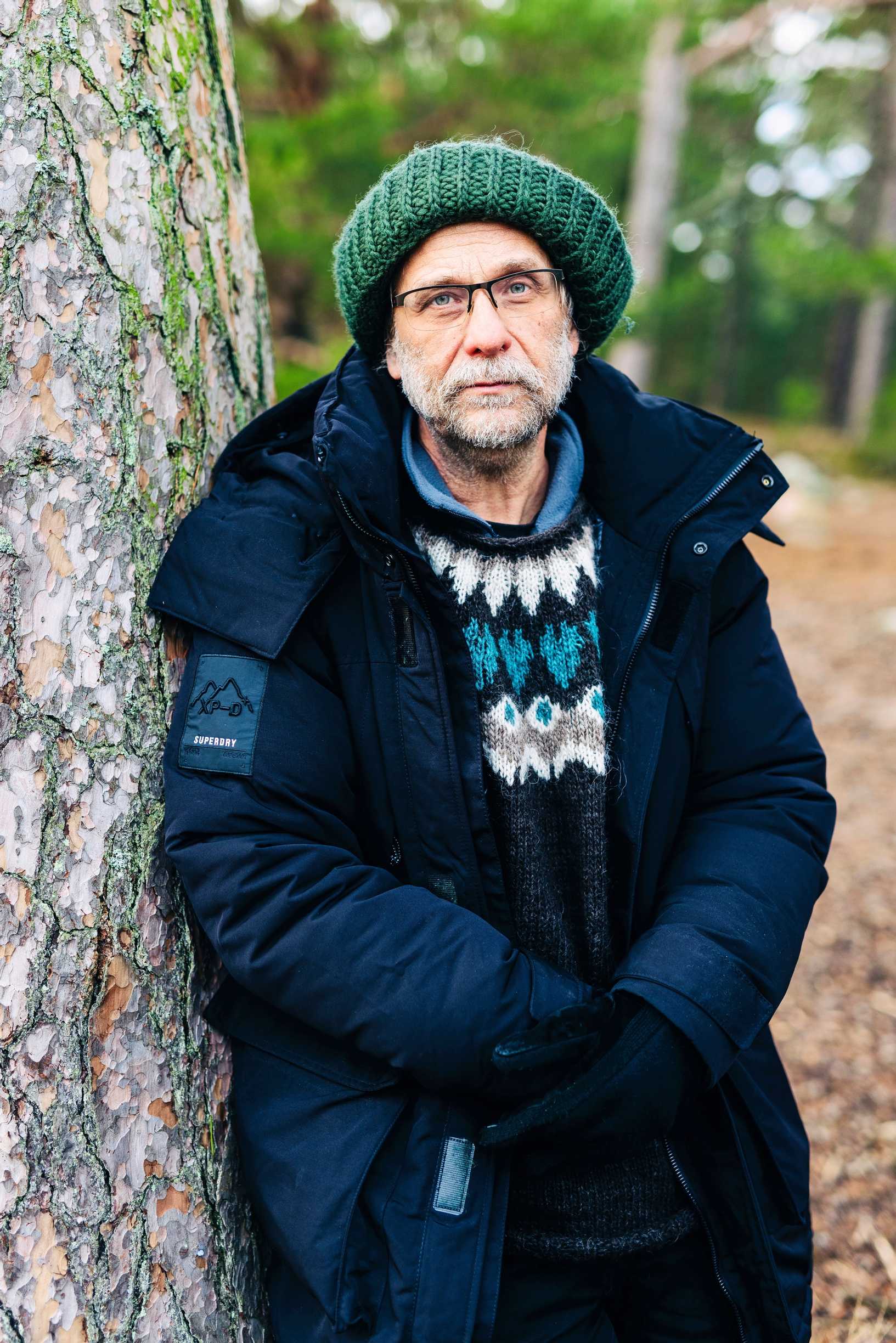 Risto Isomäki seisoo ja nojaa puunrunkoon