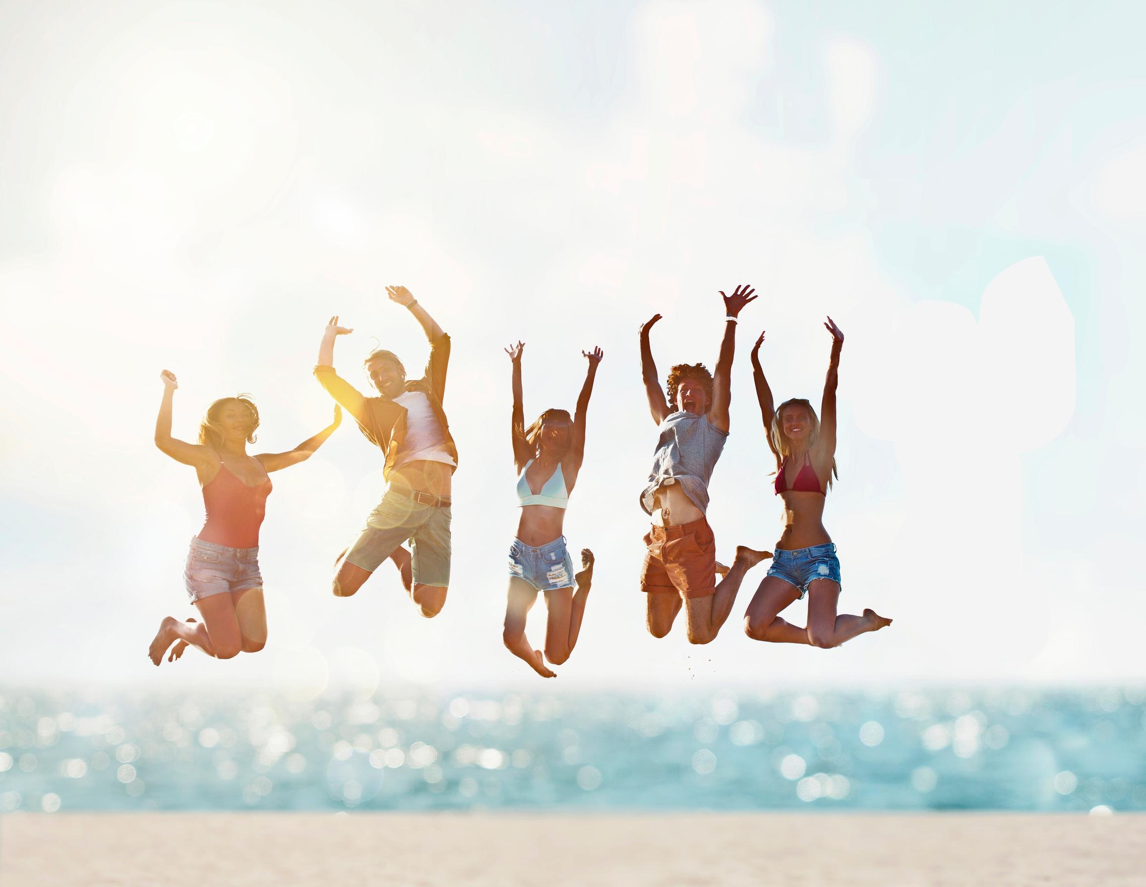 Viisi ihmistä hyppää ilmaan rantamaisemassa kädet kohotettuina ylös.